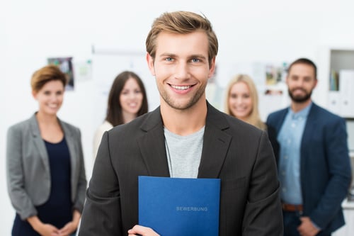 Successful smiling young male job applicant holding a blue file with his resume posing in front of his new work colleagues.