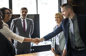 Group of businesspeople standing around a desk, two are shaking hands.