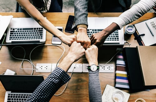 A team of five people sitting at a table with laptops group fist bumping. 