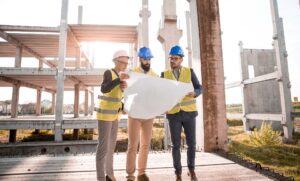 two men and a woman wearing hard hats stand on concrete in a half-finished job site, looking at architectural plans,
