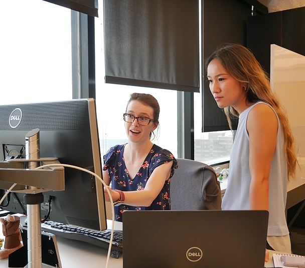 Two women discussing something in front of a computer screen; a window behind them looks out over other buildings.