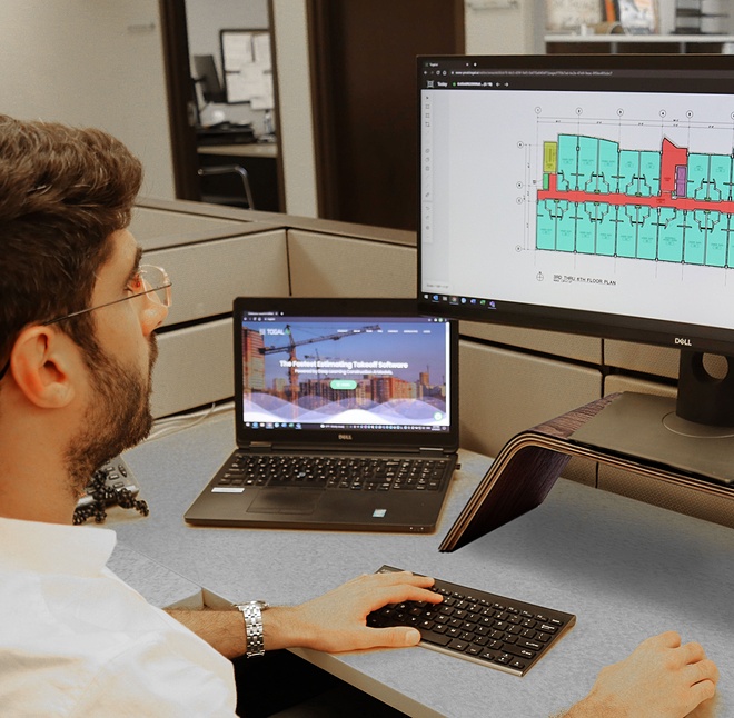 A man sits at a desk with a laptop and a large monitor, reviewing building plans. 